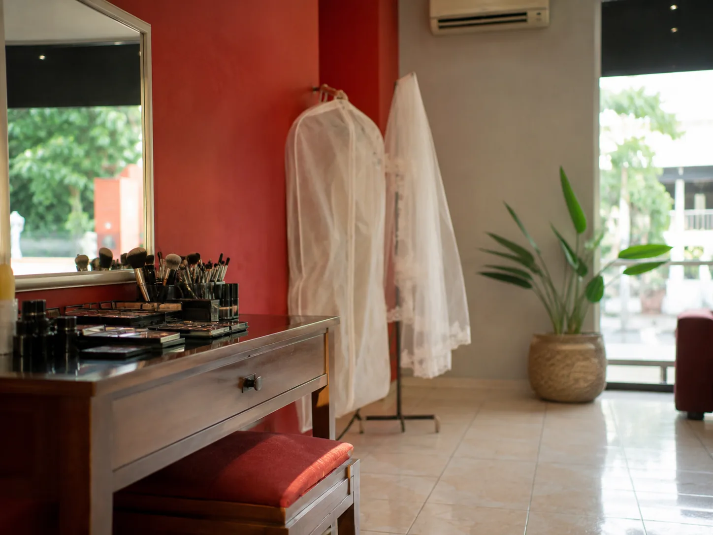 Meja rias bridal studio dengan kuas makeup dan palet, serta gaun pengantin dan veil tergantung di belakang.