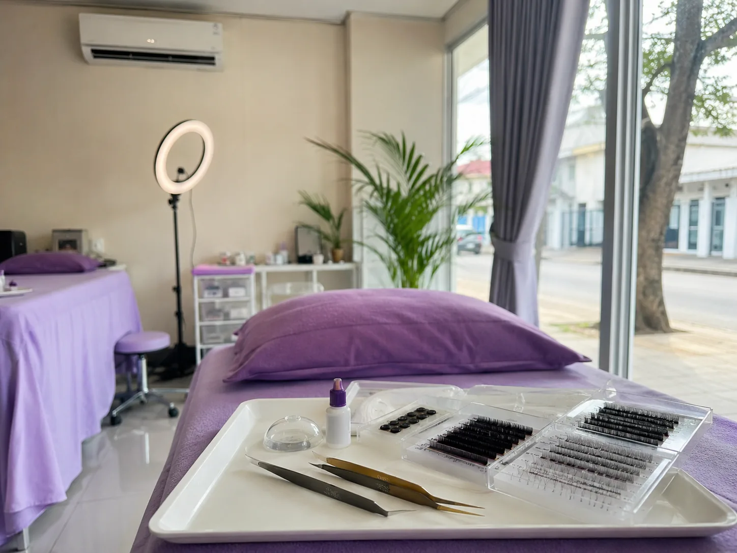 Bed perawatan eyelash extension berlapis kain ungu dengan nampan berisi pinset, lem, dan tray bulu mata, serta ring light di belakangnya.
