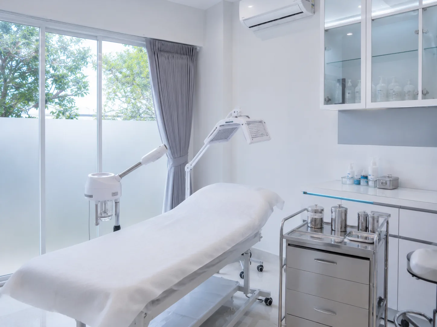 Ruang treatment klinik kecantikan serba putih dengan bed perawatan, alat LED phototherapy dan facial steamer, serta trolley stainless berisi instrumen steril.