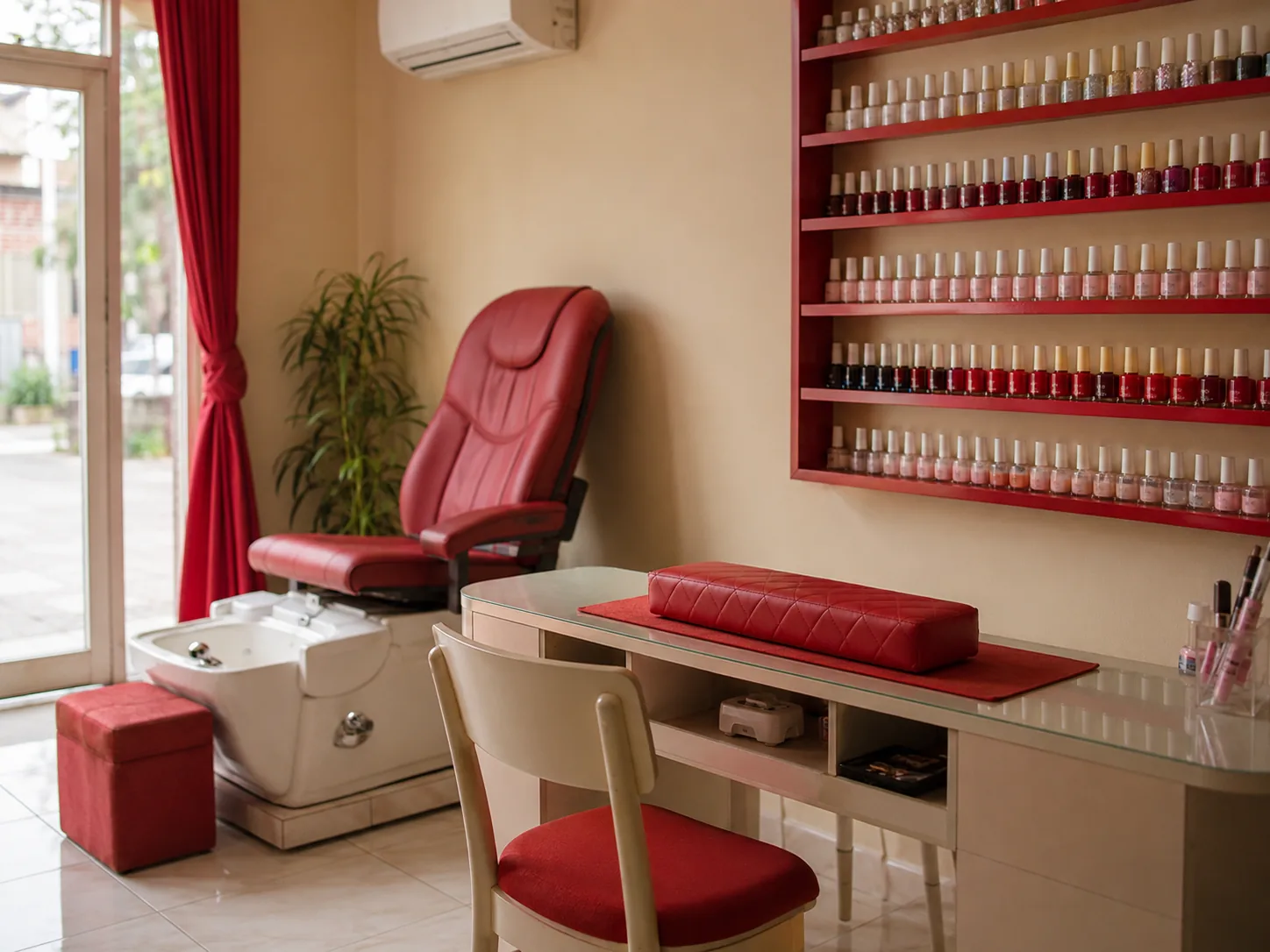 Meja manicure dengan bantalan tangan, kursi pedicure, dan rak cat kuku menempel di dinding.
