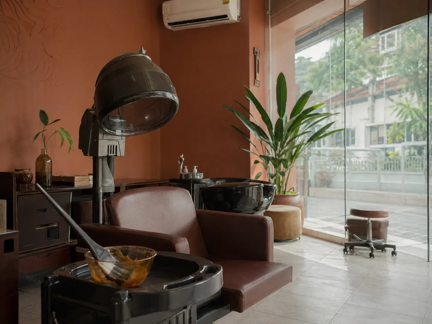 Kursi perawatan dengan hair steamer, mangkuk campuran dan kuas aplikator di sampingnya, serta wastafel keramas di ruang berdinding terakota.