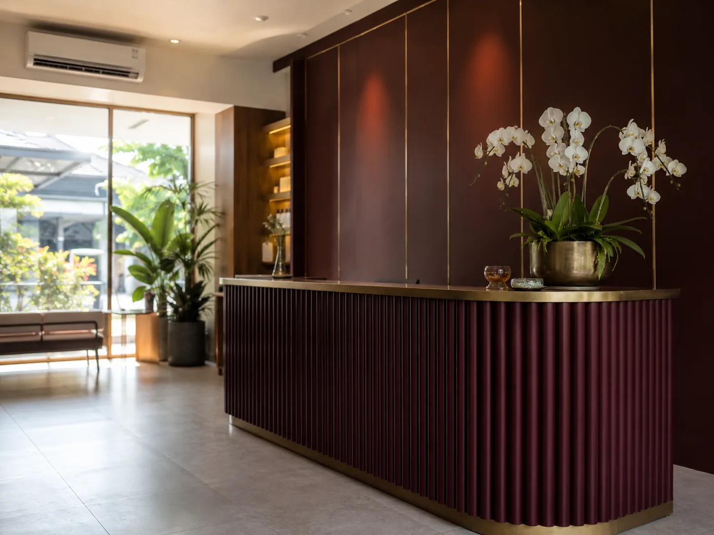 Meja resepsionis salon kecantikan premium berwarna marun dengan detail kuningan dan rangkaian anggrek putih di atasnya.