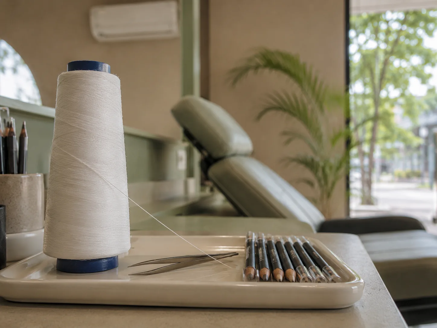 Gulungan benang threading dengan benang terurai di atas nampan, bersama pinset dan deretan pensil alis, dengan kursi perawatan alis di belakangnya.