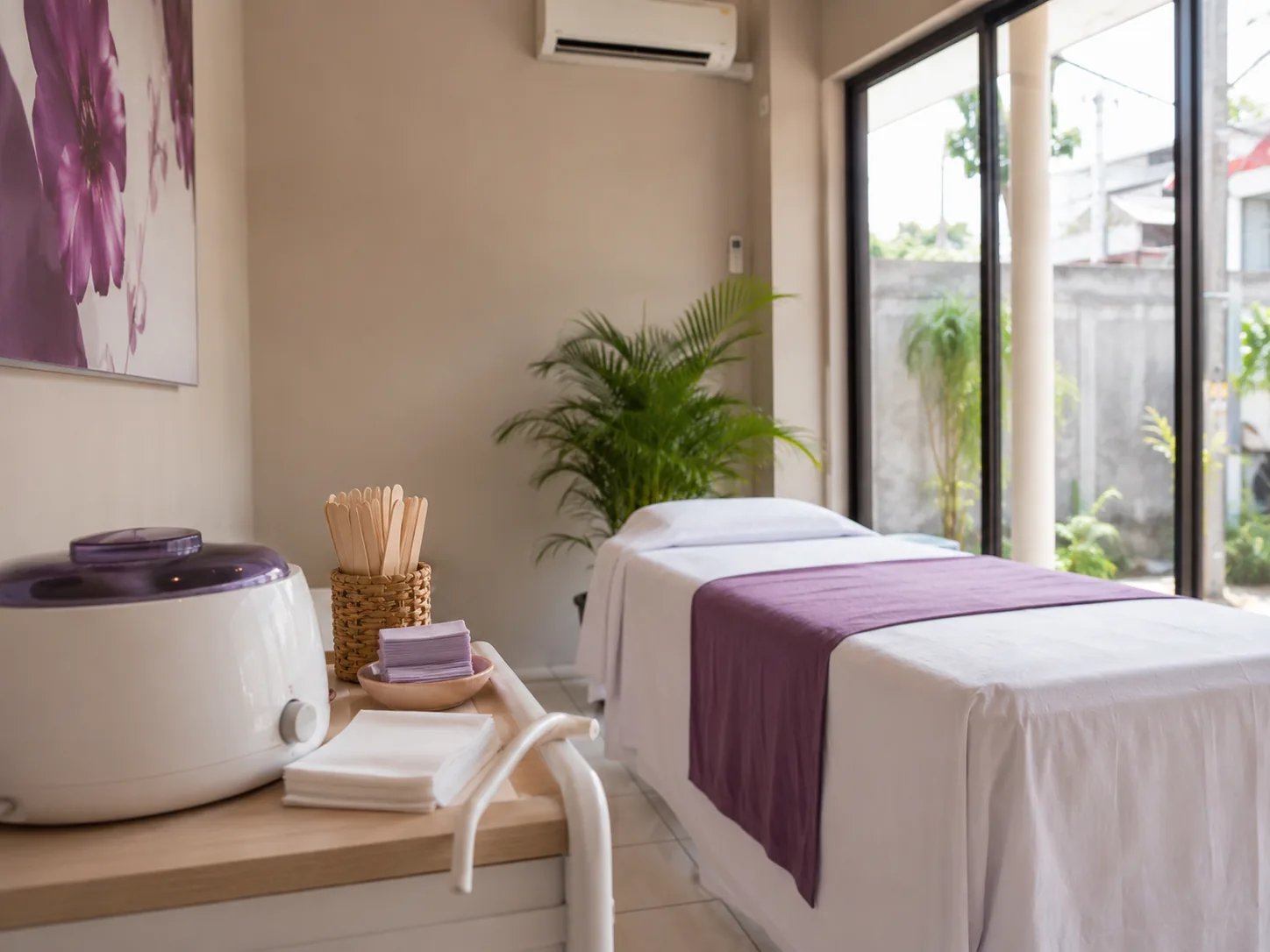 Ruang waxing dengan pemanas wax, spatula kayu dalam wadah anyaman, dan bed perawatan berlapis kain putih dengan aksen ungu.